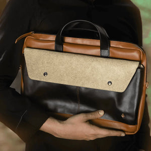 Hand holding a brown and black vegan leather briefcase with a textured flap.