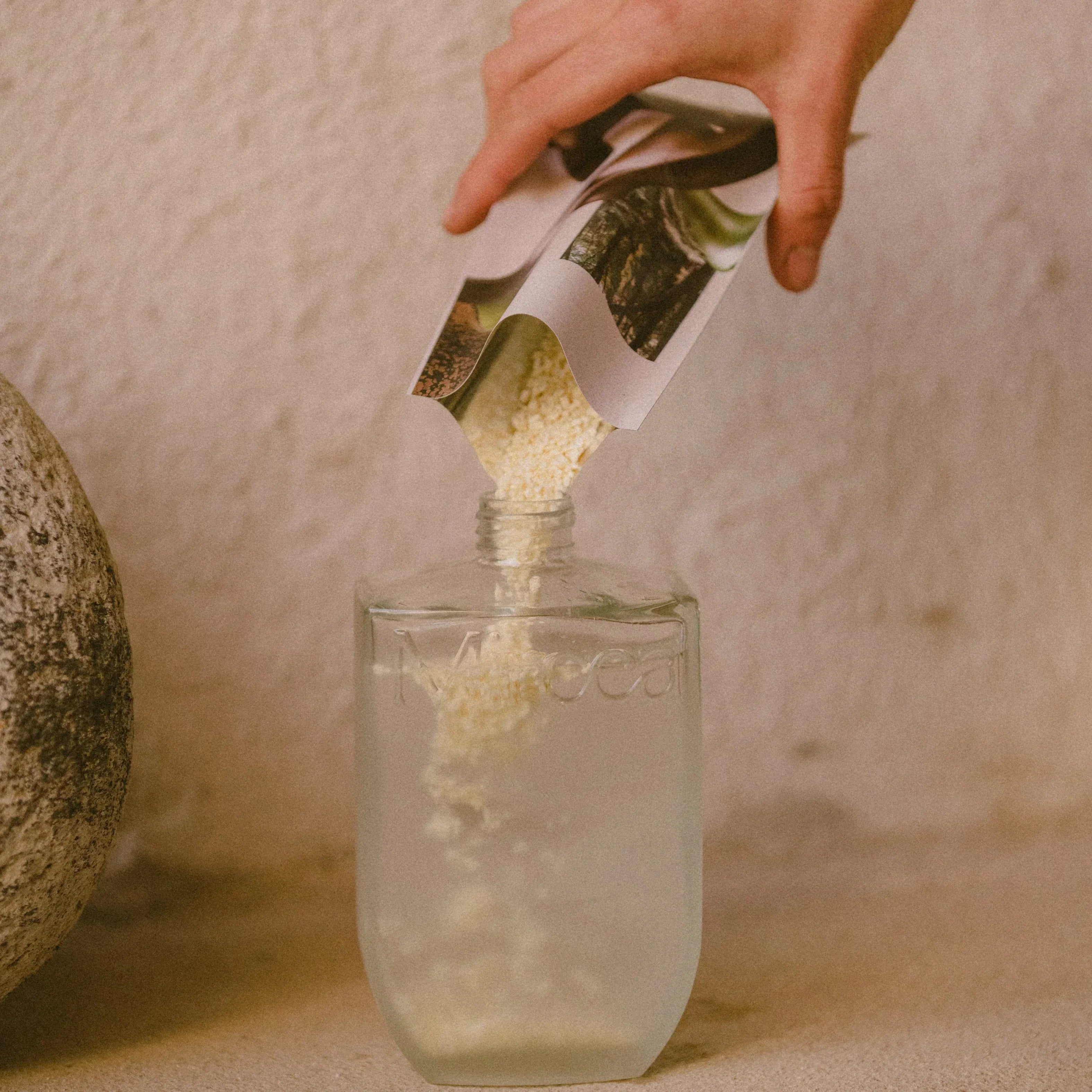 Hand pouring a green powder from a labeled container into a infinitely refillable glass bottle of water on a neutral background