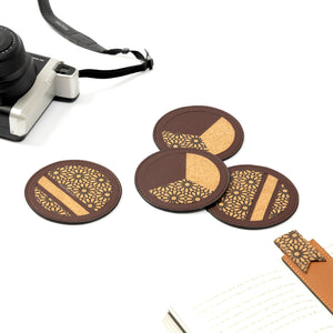 Set of four brown coasters with gold patterns on a white surface with a camera and notebook.