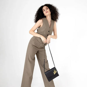 Woman in a beige sleeveless top and pants holding a black handbag with a gold emblem on a white background