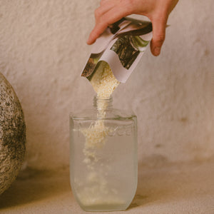 Hand pouring a green powder from a labeled container into a glass of water creating Cedar & Rosemary Hand & Bodywash on a neutral background