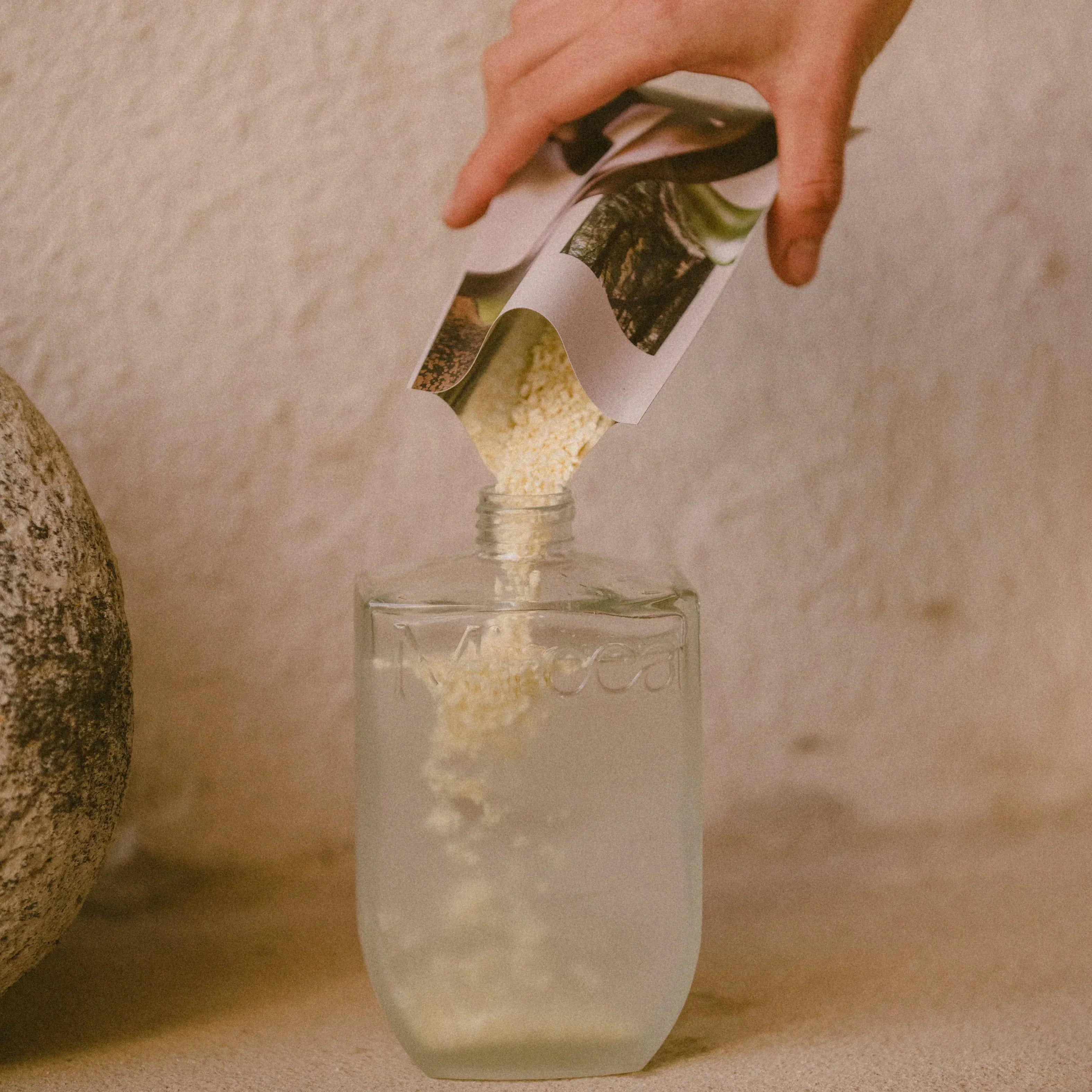 Hand pouring a green powder from a labeled container into a glass of water creating Cedar & Rosemary Hand & Bodywash on a neutral background