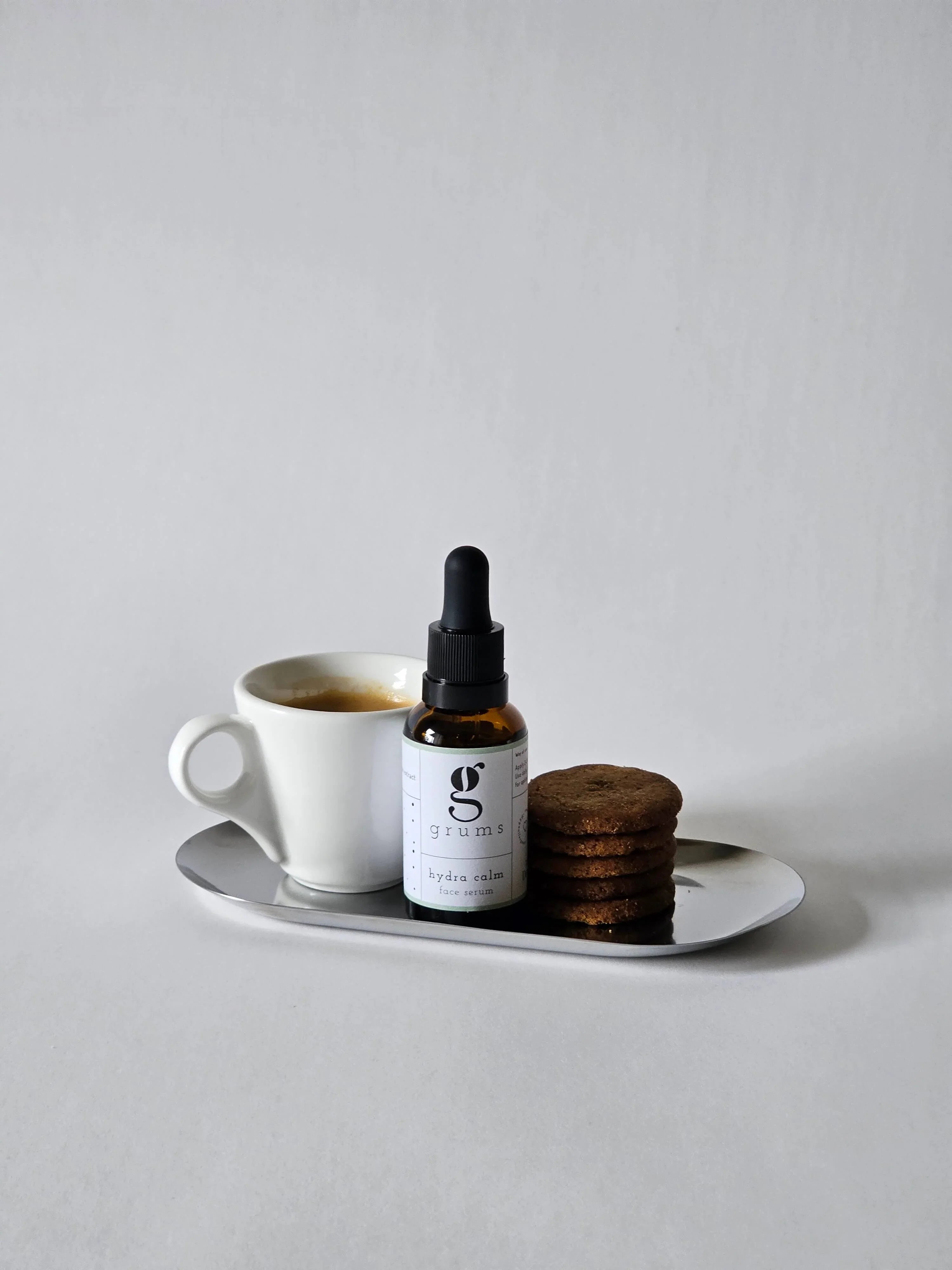 Bottle of grums hydra calm serum on a tray with a cup of coffee and cookies against a plain background