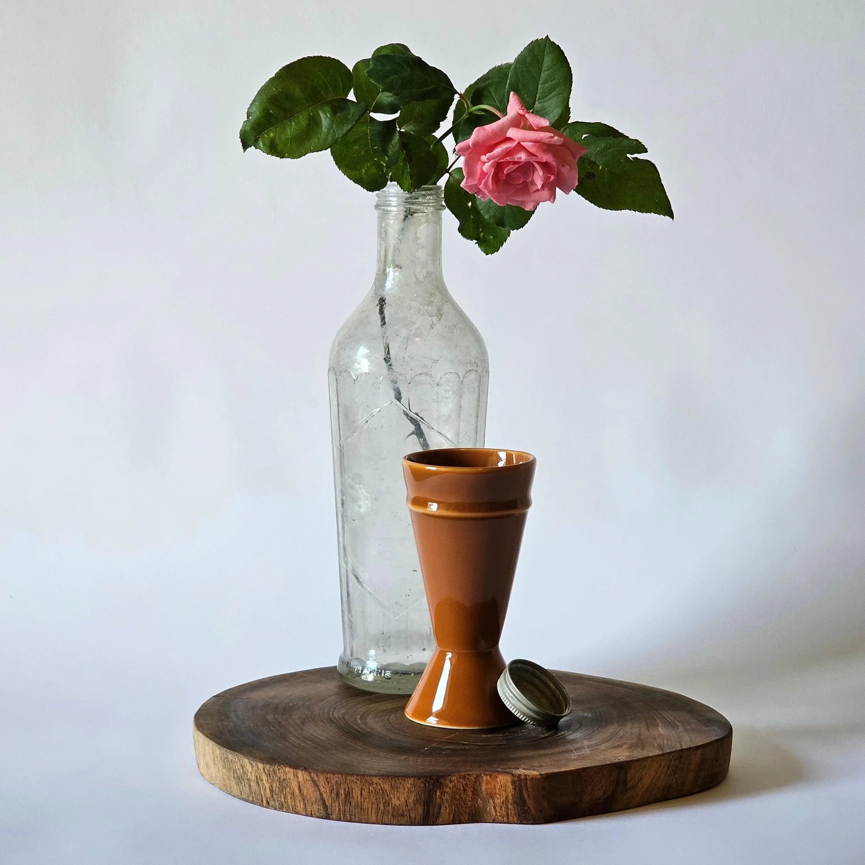 A caramel-colored Mazagran coffee cup placed on a wooden surface with a glass bottle and a pink rose in the background.