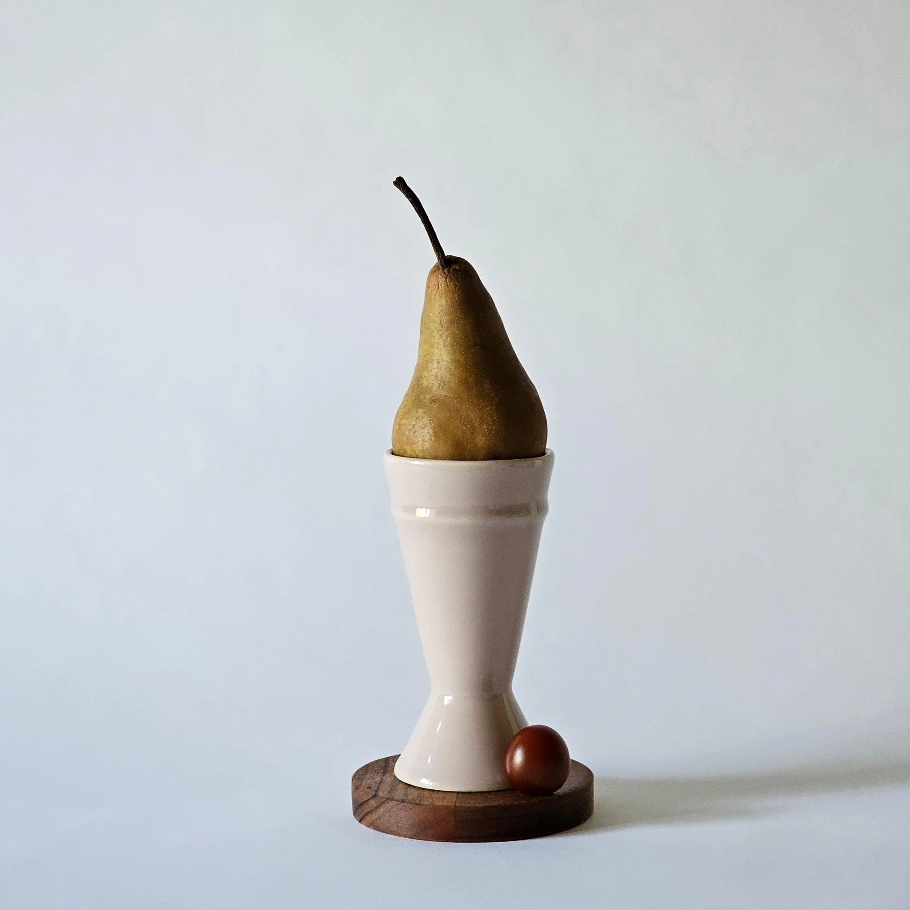 A white Mazagran coffee cup with a pink rim, featuring a pear as a playful addition to its design, displayed on a wooden stand with a small red fruit beside it.