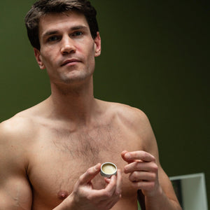 A person holding a small container of lip balm, with a mirror reflecting a green wall in the background.