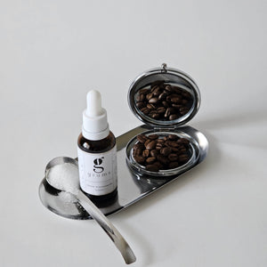 Bottle of serum with dropper, two small bowls of coffee beans, and a spoon on a white surface.