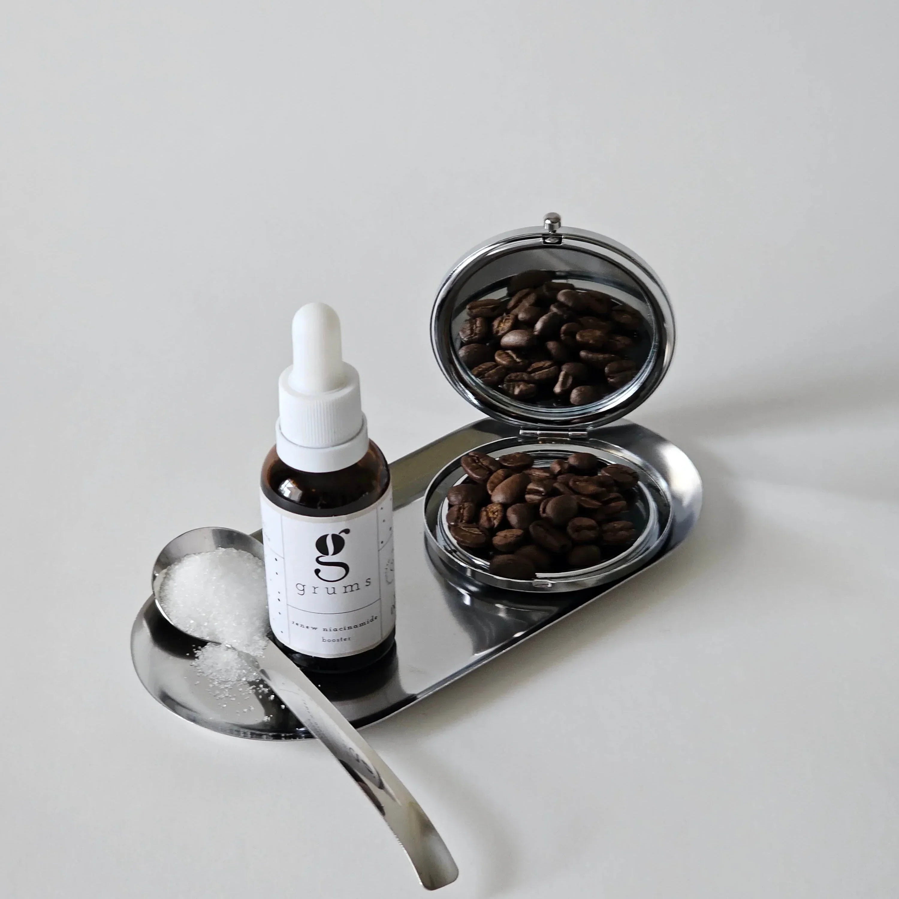 Bottle of serum with dropper, two small bowls of coffee beans, and a spoon on a white surface.