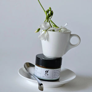 White cup with flowers, jar, and spoon on a gray background. grums hydra calm face cream balanced in the centre