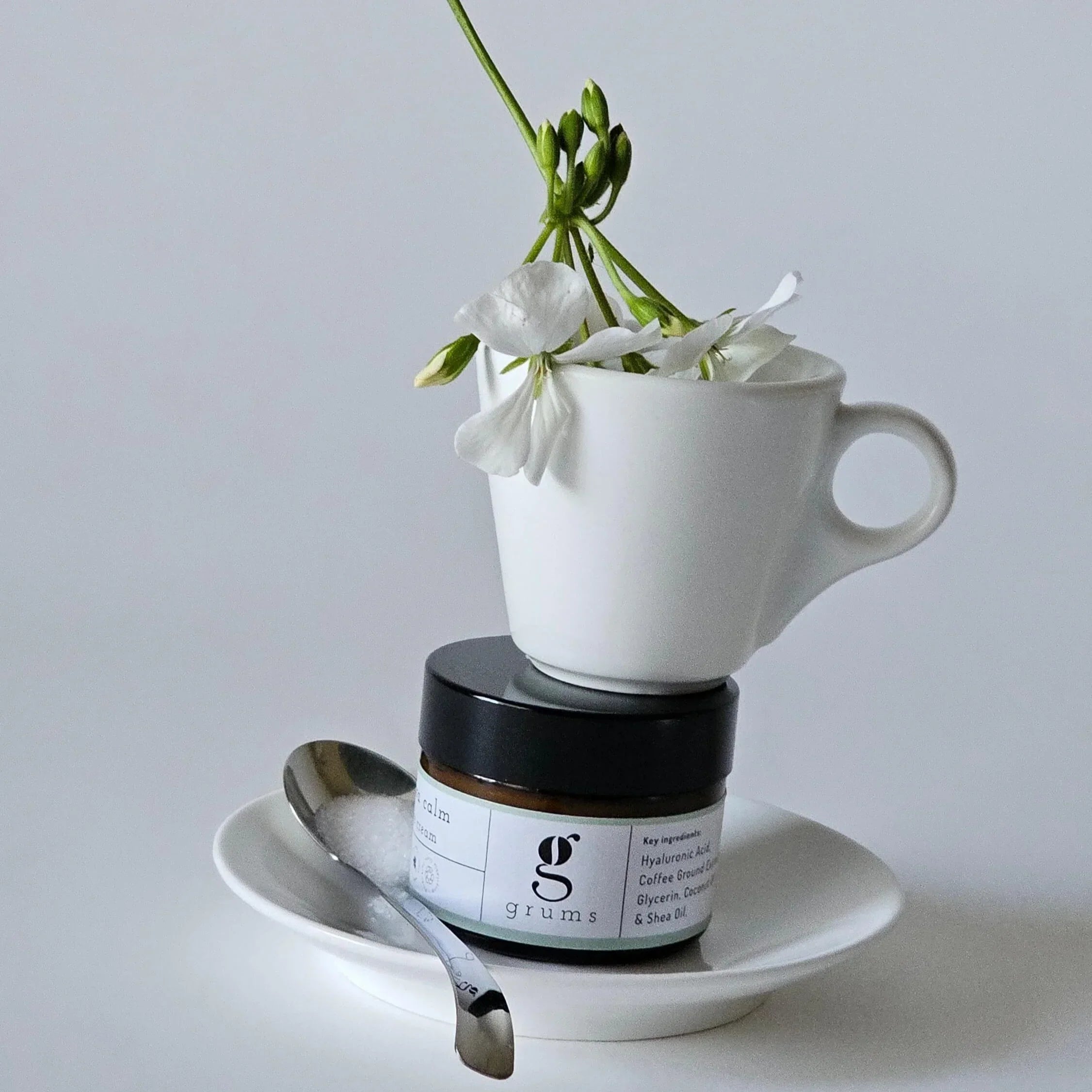 White cup with flowers, jar, and spoon on a gray background. grums hydra calm face cream balanced in the centre