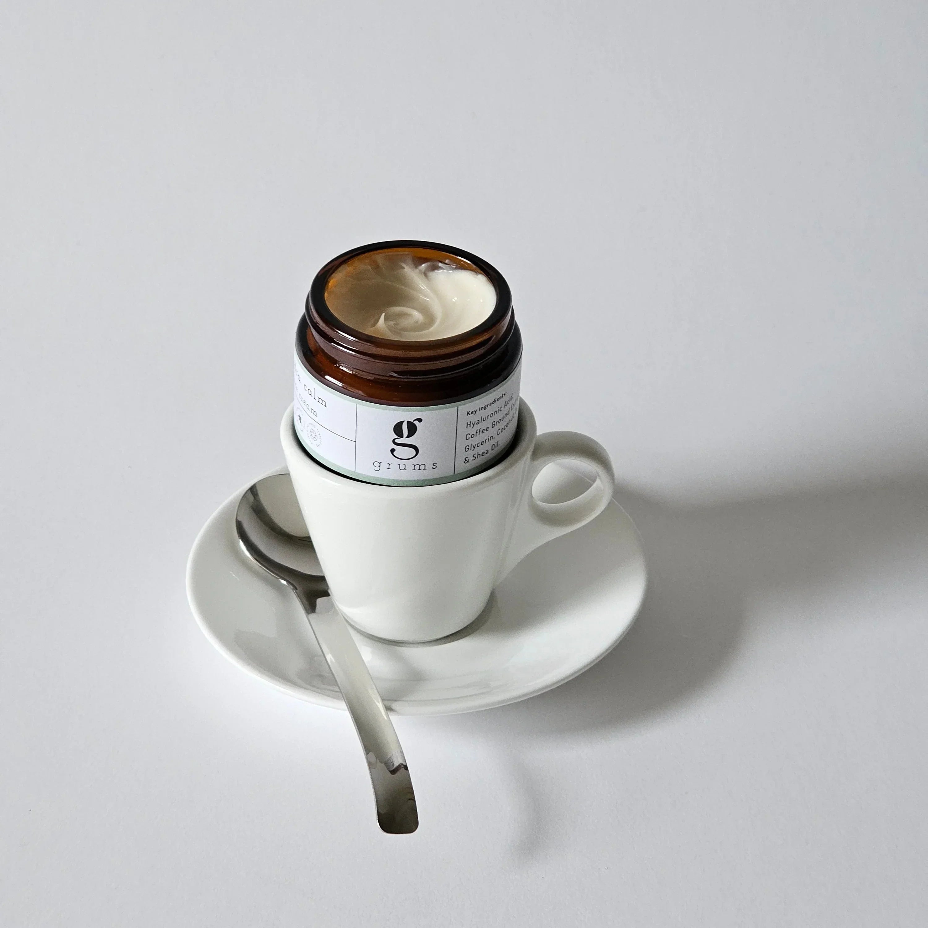grums hydra calm face cream with the lid off. the jar is sitting in a empty coffee cup with saucer and spoon