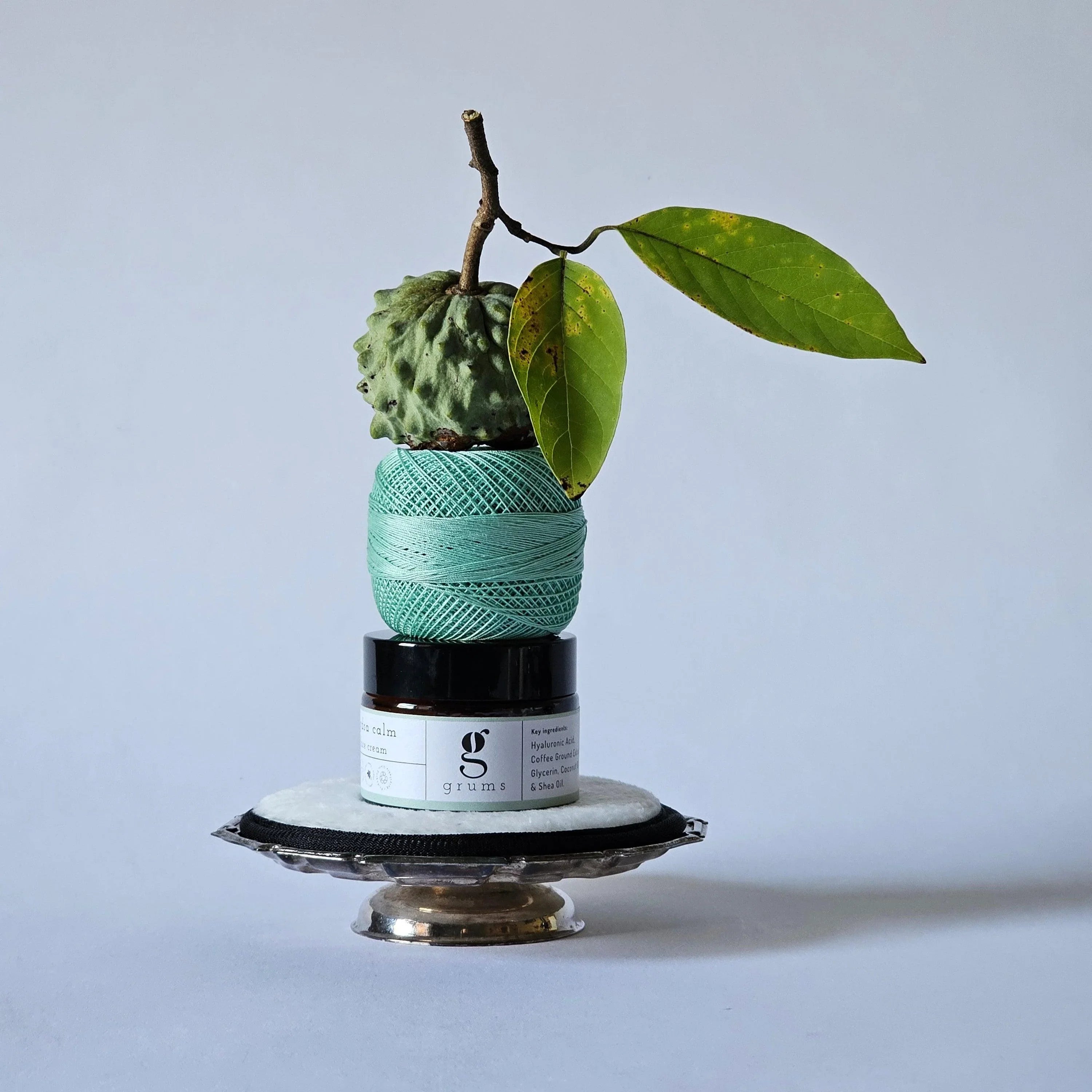 Green yarn ball with a branch and leaves on a gray background. hydra calm moisturizer sitting in the middle