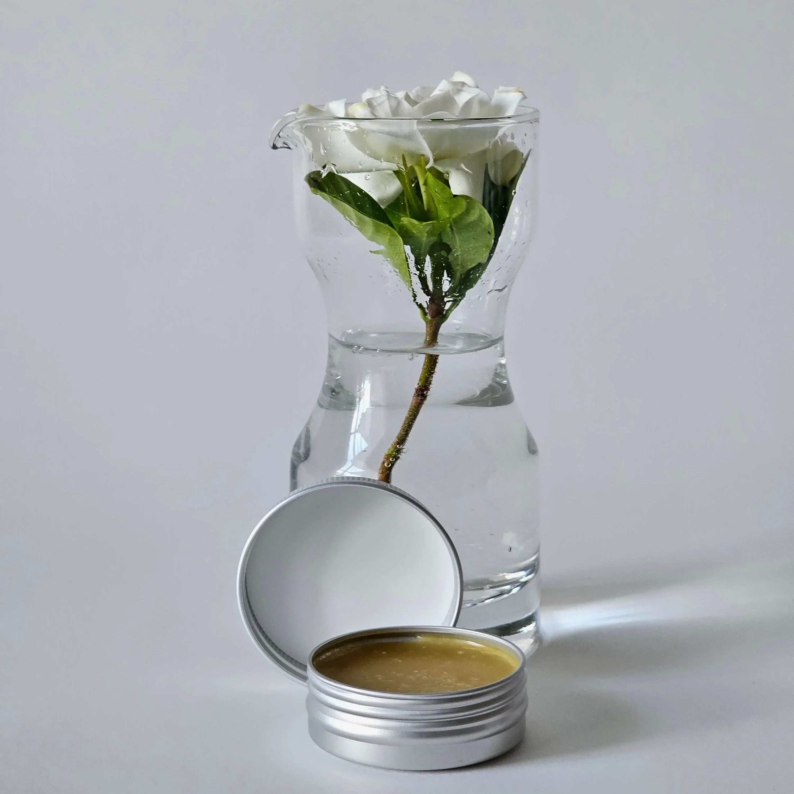 Clear glass vase with water, a branch, and a small container of grums rescue multi balm on a light gray background