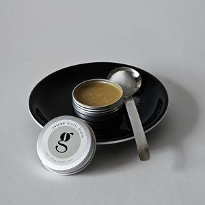 Tin of grums vegan rescue multibalm with spoon on a black plate against a gray background