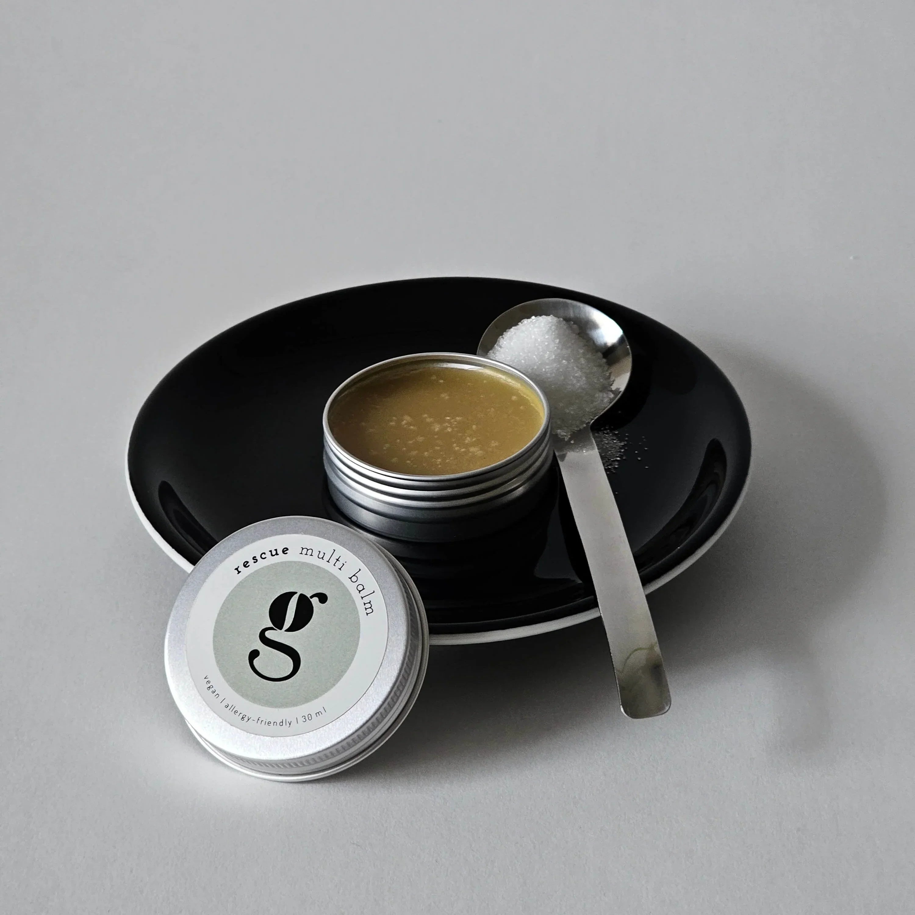 Tin of grums vegan rescue multibalm with spoon on a black plate against a gray background