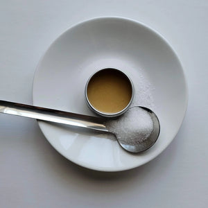 White bowl with a measuring cup and spoon on a light gray background, grums lip balm sitting in the centre