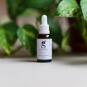 Bottle of 'grums' serum with a dropper on a white surface with green leaves in the background