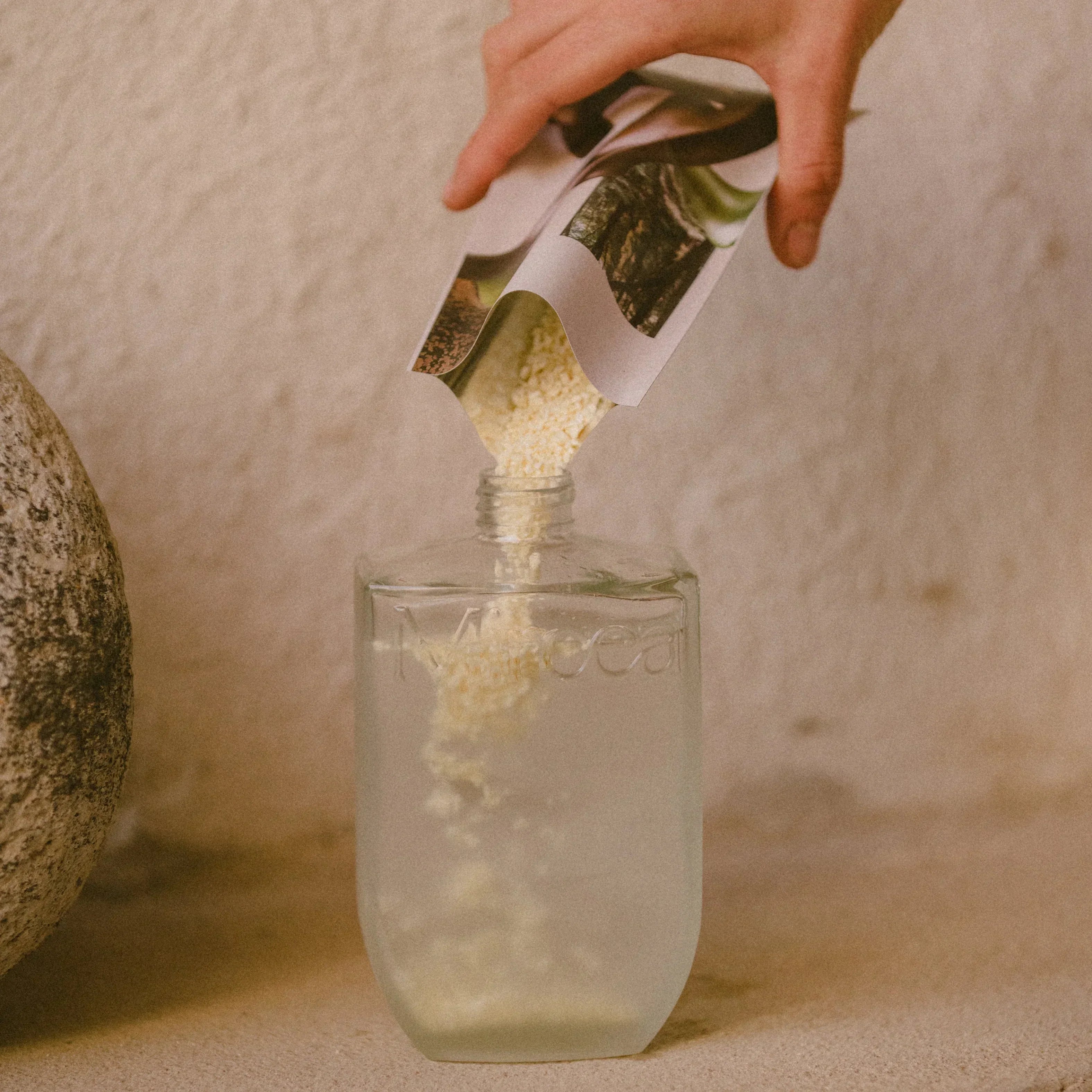 Hand pouring a green powder from a labeled container into a glass of water on a neutral background