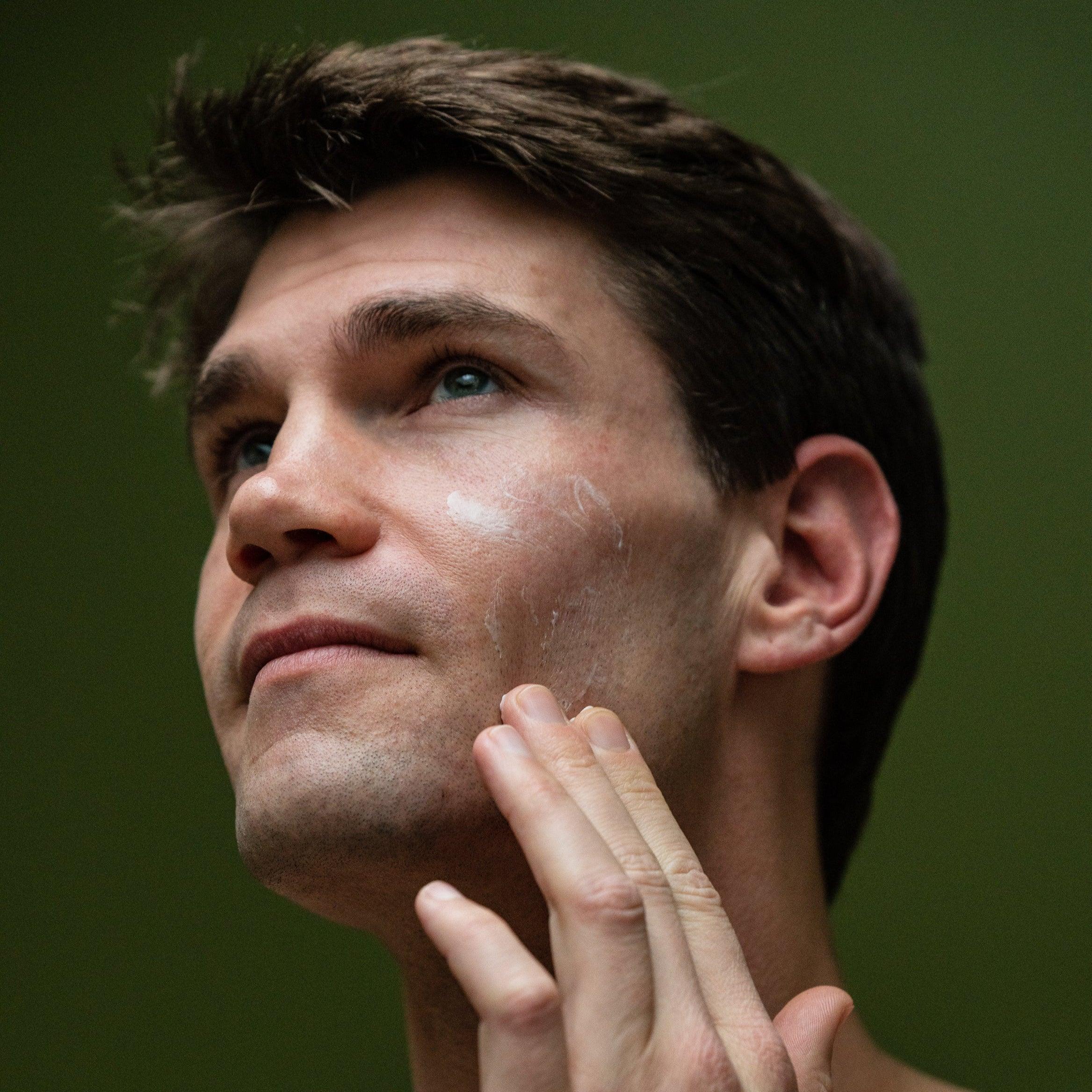 A person with short brown hair is shown applying face cream to their cheek with their finger. The background is green.