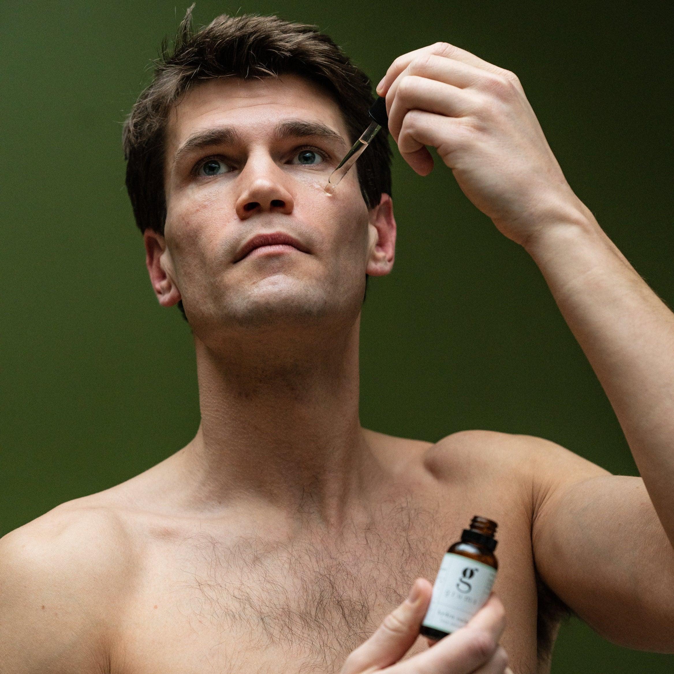 A person with short brown hair is applying face serum to their face using a dropper bottle.
