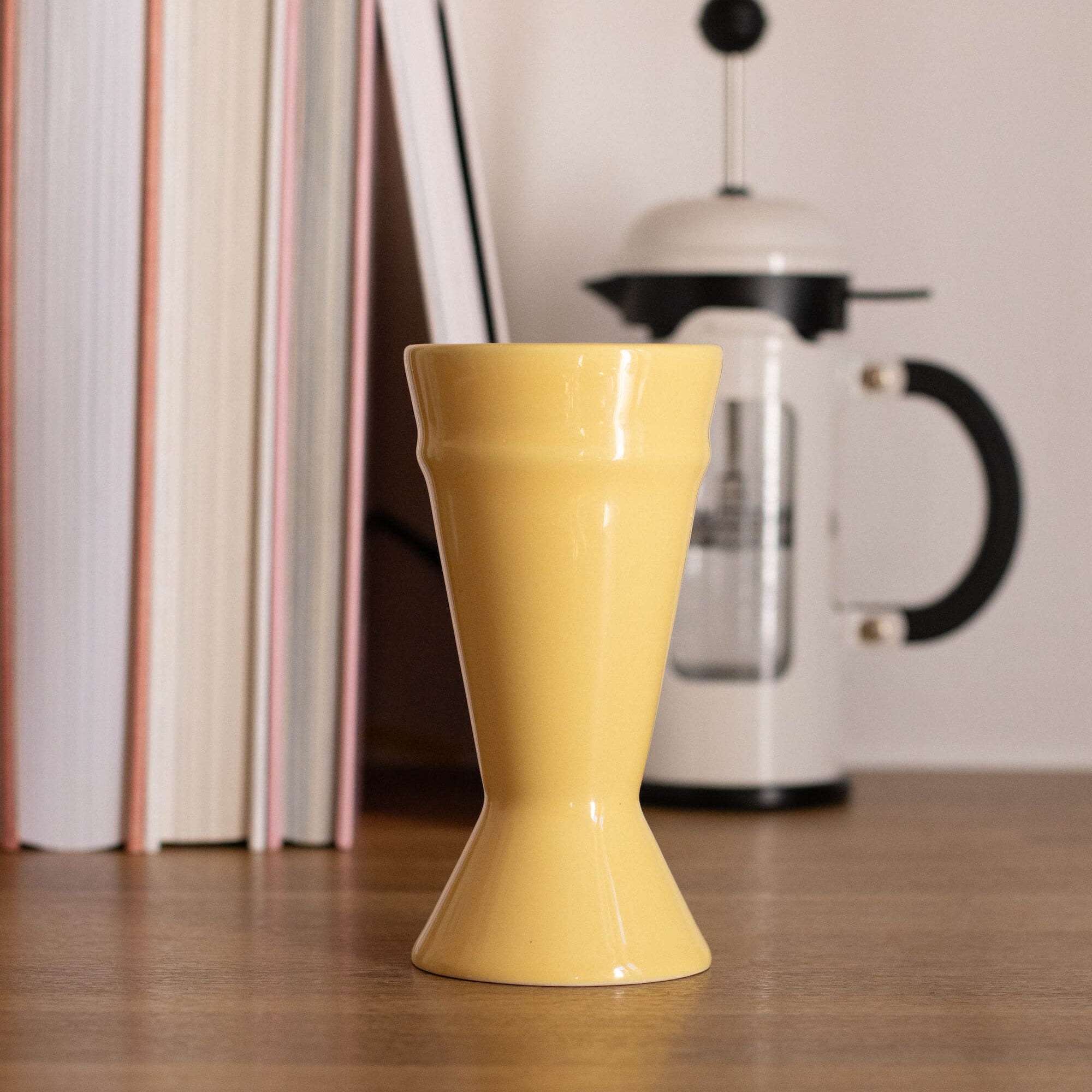 Yellow ceramic Mazagran cup on a wooden surface with books and a coffee press in the background.