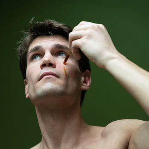 A person with a bare chest is applying a drop of coffee oil to their cheek using a dropper bottle.