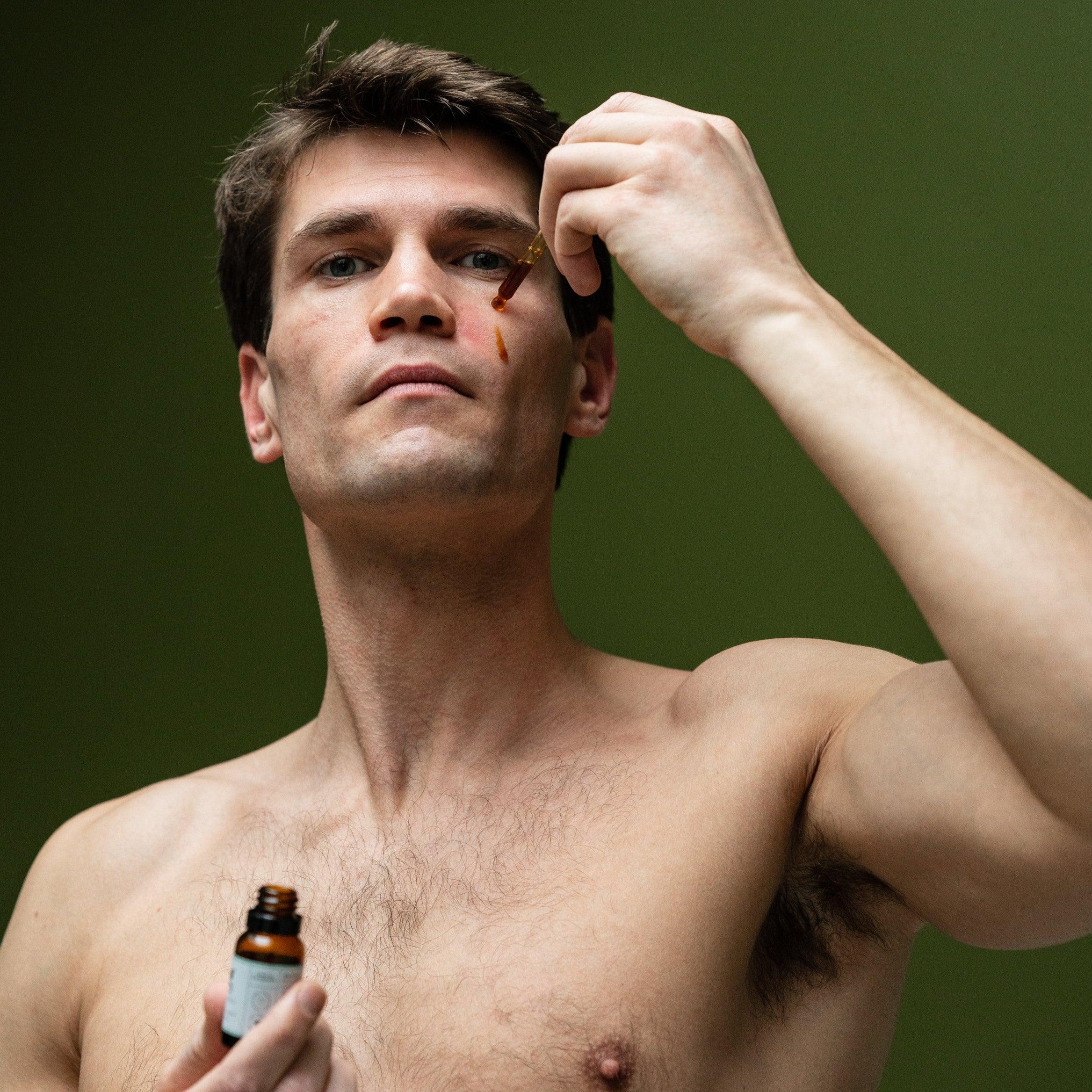 A person with a bare chest is applying a drop of coffee oil to their cheek using a dropper bottle.