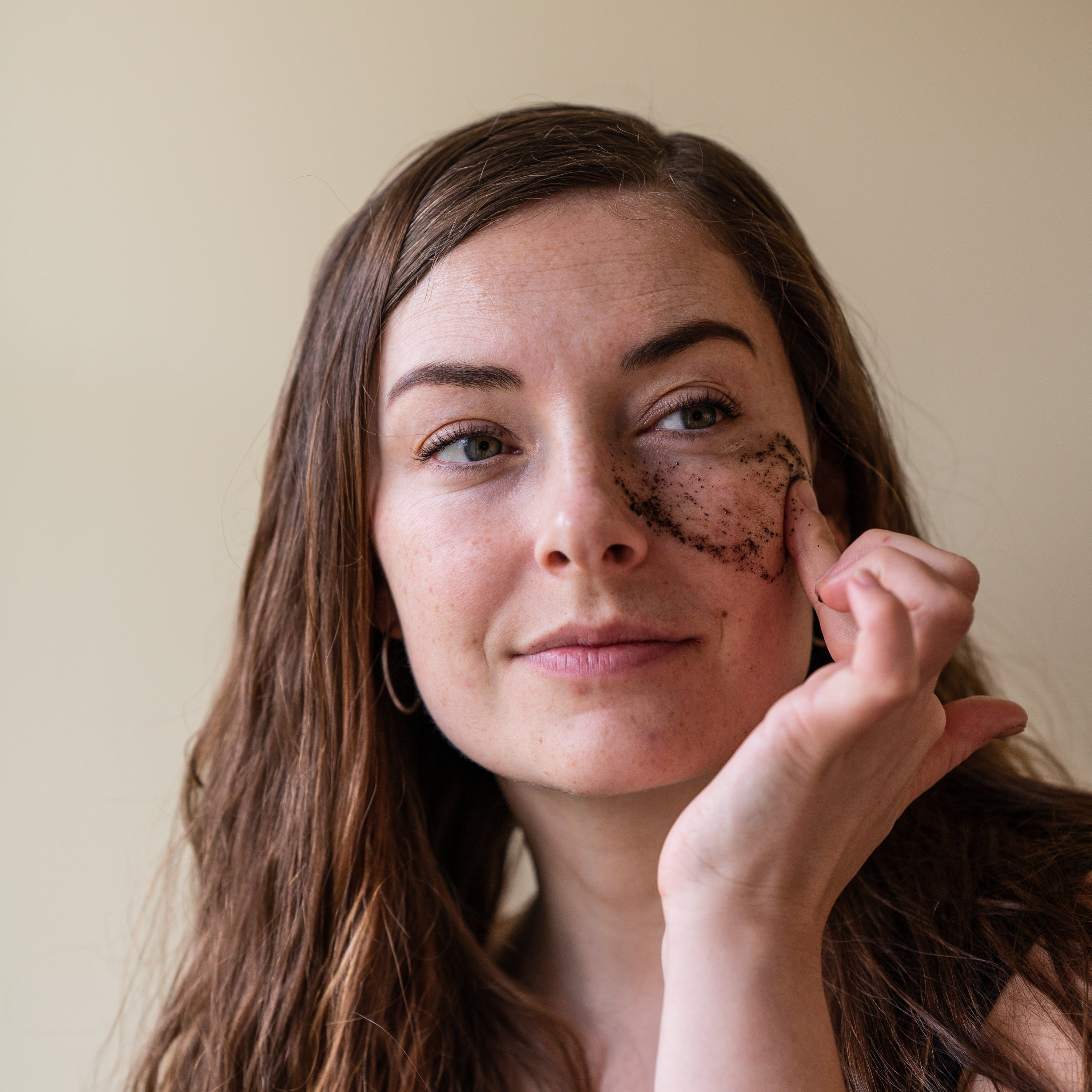Woman with brown hair and freckles on her face, applying grums coffee based face scrub & mask a leaf to her eye.