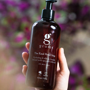 A person holding a 500ml dark plastic bottle of 'The Kind Hand Soap' by grums with a pump dispenser, label displaying the brand and product name, against a blurred background with purple flowers.