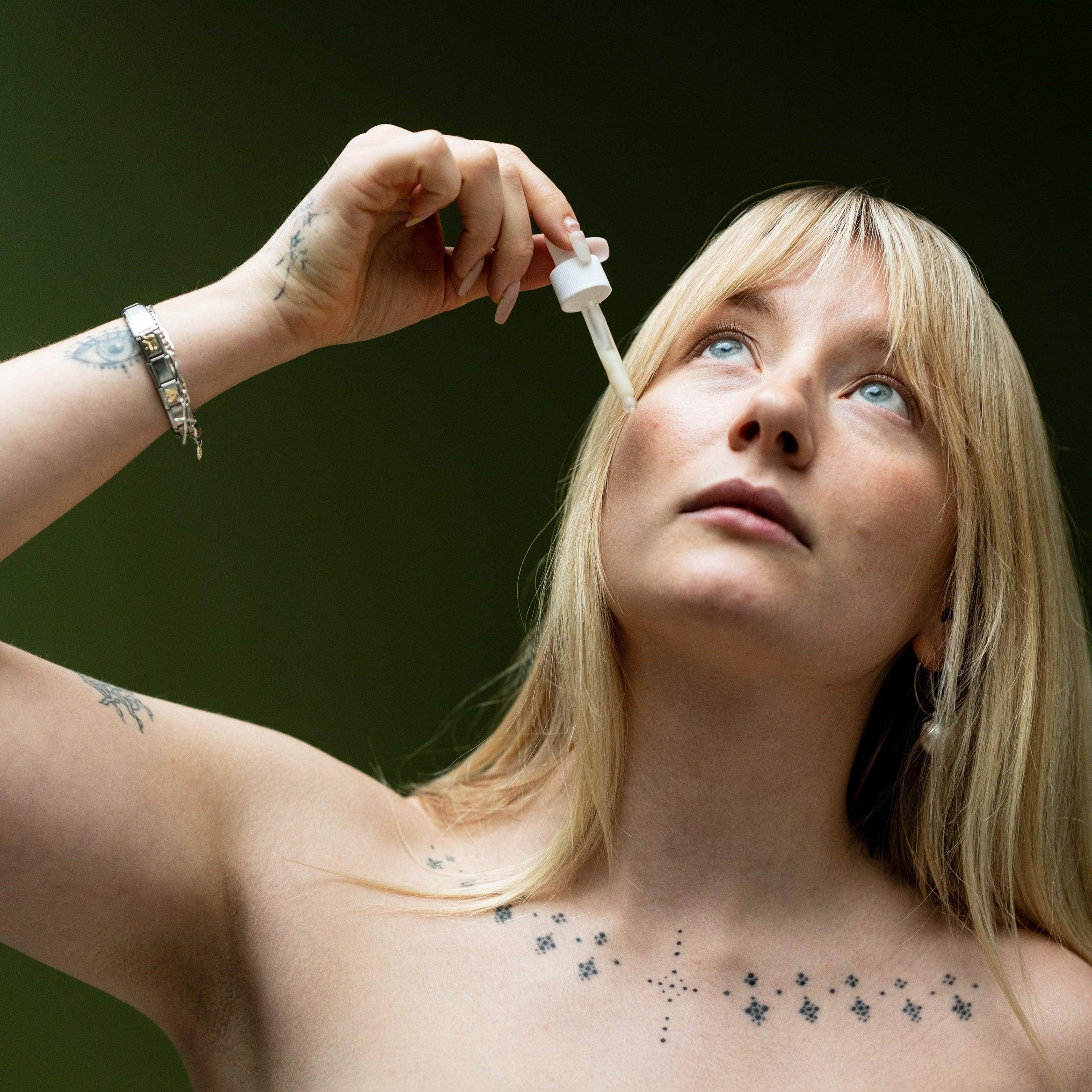 A person with light skin and blonde hair is shown holding a dropper bottle of under eye serum close to their face, looking upwards.